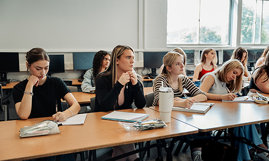 Myfyrwyr yn eistedd wrth ddesgiau ac yn ysgrifennu nodiadau mewn llyfrau nodiadau