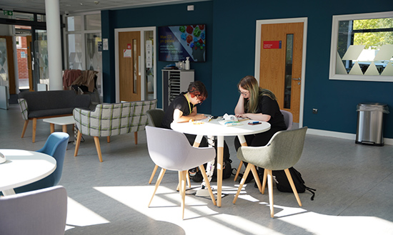 Dau berson yn eistedd wrth fwrdd mewn ardal gymunedol