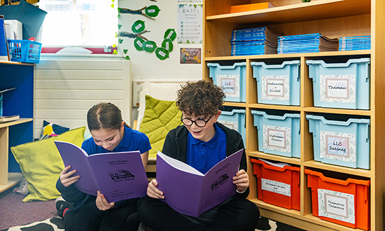 Dau ddisgybl yn eistedd ar lawr yr ystafell ddosbarth yn darllen llyfrau