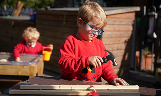 Bachgen ifanc yn chwarae y tu allan gyda morthwyl, gyda phlentyn arall yn y cefndir