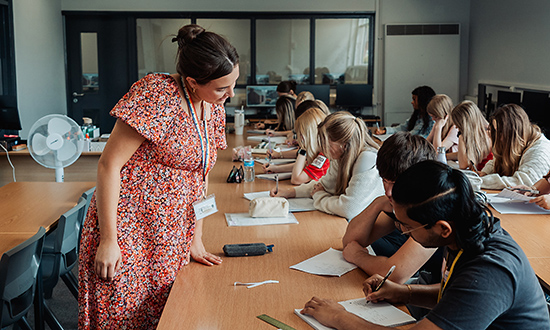 Athro yn goruchwylio gwaith disgyblion mewn ystafell ddosbarth o fyfyrwyr yn eistedd wrth ddesgiau