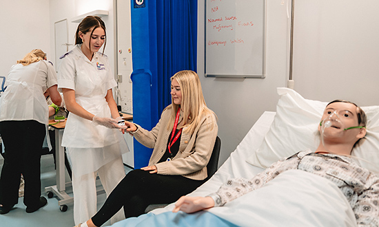 Menyw ifanc yn defnyddio offer meddygol ar fenyw arall, gyda dymi hyfforddi mewn gwely yn y blaendir